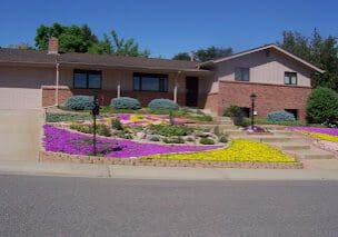 A brick house with a vibrant, colorful flower garden in the front yard under a clear blue sky.